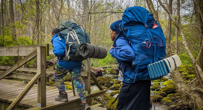 two young people wearing backpacks cross a creek over a bridge on a backpacking trip